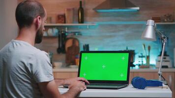 Businessman looking at green screen monitor sitting at home in kitchen. Freelancer watching desktop monitor display with green mockup, chroma key, during night time working overtime. video