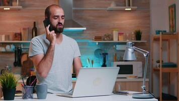 aburrido hombre en camiseta bostezando mientras hablando en teléfono inteligente trabajando a hogar con ordenador portátil. ocupado enfocado empleado utilizando moderno tecnología red inalámbrico haciendo tiempo extraordinario para trabajo leyendo escribiendo, buscando video