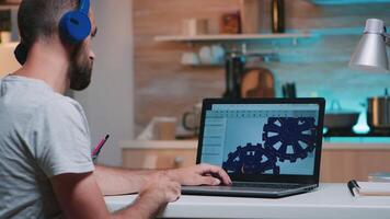 Man architect with wireless headset using laptop while working at home at night sitting in the kitchen. Industrial employee studying prototype idea on personal computer showing cad software on display video