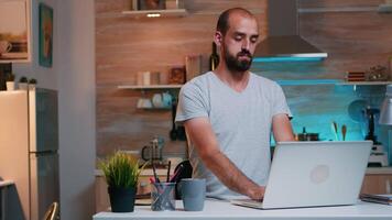 cansado hombre estirar después largo trabajando remotamente día desde hogar sentado en moderno cocina, trabajo excesivo. ocupado enfocado empleado utilizando moderno tecnología red inalámbrico haciendo tiempo extraordinario para trabajo leyendo escritura video