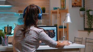 Architect with wireless headset using laptop while working at home at night sitting in the kitchen. Industrial female engineer studying on personal computer showing cad software. video