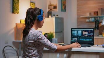 Woman editor with headset working with footage and sound sitting in home kitchen. Woman videographer editing audio film montage on professional laptop sitting on desk in midnight video