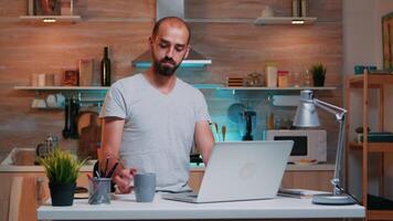 pigiste en buvant café travail de Accueil en retard à nuit en utilisant portable dans moderne cuisine. concentré employé en utilisant moderne La technologie réseau sans fil Faire heures supplémentaires pour emploi en train de lire en écrivant, recherche video