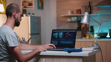 Professional videographer working in editing app wearing headphones in front of laptop sitting in home kitchen. Freelancer processing audio film montage on professional laptop in midnight video