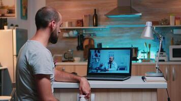 Doctor videoconferencing remote patient consulting at night, man listening sitting in front of laptop in the kitchen. Sick man discussing during conference about symptoms holding bottle of pills video