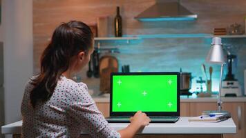 Business woman looking at green screen laptop sitting at home in kitchen. Freelancer watching desktop monitor display with green mockup, chroma key, during night time working overtime. video
