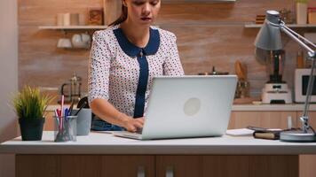 o negócio mulher trabalhando a partir de casa sentado dentro a cozinha digitando em computador portátil atrasado às noite. ocupado focado empregado usando moderno tecnologia rede sem fio fazendo hora extra para trabalho lendo escrita, procurando video