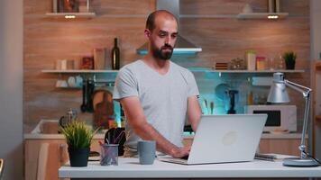 cansado soñoliento hombre bostezando, trabajando a hogar escritorio en el cocina. ocupado trabajo excesivo enfocado empleado utilizando moderno tecnología red inalámbrico haciendo tiempo extraordinario para trabajo leyendo escribiendo, buscando video