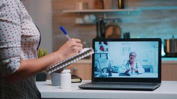 dame schrijven aantekeningen gedurende online medisch overleg luisteren vrouw dokter zittend in voorkant van laptop in de keuken. ziek persoon bespreken gedurende conferentie over symptomen en behandeling. video