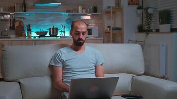 Focused man browsing information on network using laptop computer sitting in front of television. Caucaisan male lying on sofa late at night in kitchen using modern technology wireless video