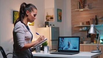 mujer Hablando en línea con médico tomando notas durante tele salud sentado en hogar cocina. enfermo dama que se discute durante virtual consulta acerca de síntomas participación cuaderno y escritura tratamiento video