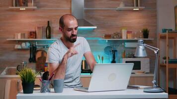 controlo remoto homem empreendedor tendo conferência com colegas usando computador portátil sentado dentro a cozinha atrasado às noite. ocupado empregado usando moderno tecnologia rede sem fio lendo escrita, procurando video