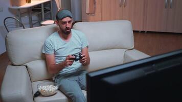 Upset pro gamer losing videogamer competition using wireless joystick during gaming competition. Disappointed player sitting in front of television on sofa making loser gesture late at night video