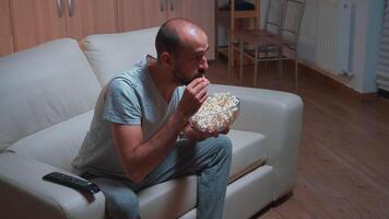 geconcentreerd Mens chillen Aan sofa in voorkant van televisie aan het kijken film serie terwijl aan het eten popcorn. Kaukasisch mannetje in pyjama op zoek Bij vermaak shows laat Bij nacht in keuken video