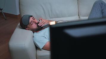 caucasico maschio con occhio dormire maschera mangiare Popcorn mentre Guardando divertimento tv mostrare. stanco uomo seduta nel davanti di televisione durante film film in ritardo a notte nel cucina video