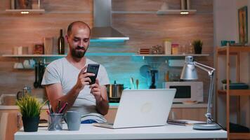 trabalhador autonomo levando pausa e procurando em telefone sentado em cozinha mesa trabalhando a partir de lar. ocupado focado empregado usando moderno tecnologia rede sem fio fazendo hora extra para trabalho lendo escrevendo video