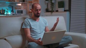 Focused man sitting on sofa in pajamas while talking with collegue during videocall conference using laptop computer. Caucasian male working at communication project late at night in kitchen video