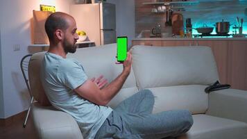 konzentriert Mann reden mit seine freunde auf Smartphone mit spotten oben Grün Bildschirm Chroma Schlüssel Anzeige. kaukasisch männlich mit modern Technologie kabellos während Sitzung auf Sofa spät beim Nacht im Küche video
