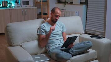 Focused man with beard sitting on couch in front of television while talking with teammates during online meeting videocall use tablet computer. Caucasian male using modern technology wireless video