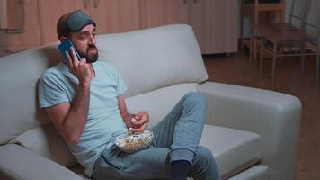 Caucasian male resting on couch eating popcorn while talking on phone with friend about social network. Relasex man sitting in front of television watching entertainment show late at night in kitchen video