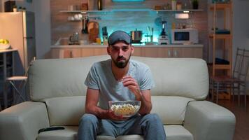 Concentrated man with sleep eye mask sitting in front of television watching lifestyle show. Caucasian male relaxing on couch holding popcorn bowl late at night in kitchen video