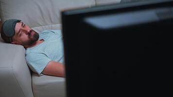 Man with eye sleep mask lying on sofa while watching entertainment movie holding popcorn bowl in hands. Caucasian male sitting in front of television during tv show late at night in kitchen video