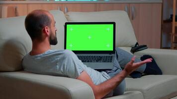 Focused man looking laptop computer with at mock up green screen chroma key display while laying on couch working at online e-learning project. Tired person using isolated pc late at night in kitchen video