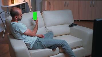 Caucasian male looking phone with at mock up green screen chroma key display while lying on sofa browsing on internet. Caucasian male using modern technology wireless late at night in kitchen video