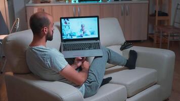 Doctor consulting sick patient during coronavirus quarantine using laptop computer for videocall consultation sitting on sofa. Caucasian man disscusing about illness symptoms and headlcare treatment video