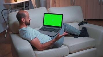Tired man sitting on sofa while browsing marketing information using laptop computer with mock up green screen chroma key display. Caucasian male using modern technology wireless late at night video