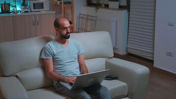 Caucasian male sitting in front of television while working at communication project before deadline on laptop. Tired man browsing on internet using network technology late at night in kitchen video