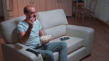 caucasico maschio seduta su divano con Popcorn ciotola nel mani mentre guardare a film serie su televisione. stanco uomo nel pigiama Guardando a divertimento Spettacoli in ritardo a notte nel cucina video