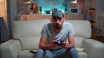 Front view of pro gamer sitting on sofa in front of television while playing videogames competition using wireless joystick. Cheerful man with sleep mask enjoying time alone late at night in kitchen video