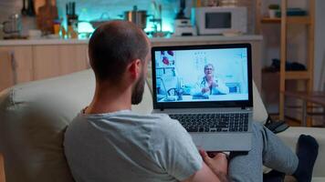 Physician doctor consulting online patient during coronavirus quarantine late at night in kitchen. Caucasian male sitting in front of laptop disscusing about illness symptoms and healthcare treatment video