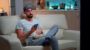 aburrido hombre sentado en sofá participación palomitas de maiz cuenco mientras mensajes de texto mensaje en social red utilizando moderno teléfono. caucásico masculino acecho televisión Deportes serie tarde a noche en cocina video