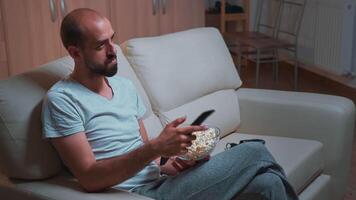 caucasico maschio chilling su divano con Popcorn ciotola nel mani mentre Guardando film serie su televisione. stanco uomo nel pigiama guardare a divertimento Spettacoli in ritardo a notte nel cucina video