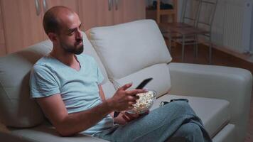 Caucasian male chilling on sofa with popcorn bowl in hands while watching movie series on television. Tired man in pajamas looking at entertainment shows late at night in kitchen video