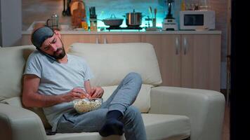 relaxado homem falando sobre Internet conexão em Smartphone. caucasiano masculino sentado em sofá assistindo entretenimento filmes enquanto comendo Pipoca atrasado às noite dentro cozinha video