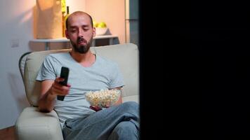 Man late at night in his apartment looking at TV entertainment, laughing and having fun while eating popcorn snack. Smiling caucasian person leisure time on the sofa in front of television video
