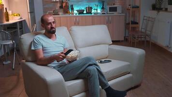 uomo con barba Guardando tv mostrare utilizzando a distanza controllo per modificare canali su televisione. caucasico maschio rilassante su divano con Popcorn ciotola nel mani guardare divertimento serie a in ritardo a notte video