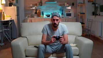 Focused man holding joystick while sitting in front of television winning online games competition. Caucasian male with sleep mask lying on couch making winner gesture late at night in kitchen video