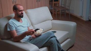 Concentrated man sitting in front of television using control remote while watching movie film. Caucasian male in pajamas looking at entertainment shows late at night in kitchen video