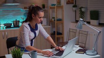 concentrado freelance mulher trabalhando hora extra para terminar uma projeto a partir de casa cozinha. ocupado focado empregado usando moderno tecnologia rede sem fio fazendo hora extra para trabalho lendo digitando, procurando video