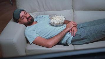 fatigué homme mensonge sur canapé en train de regarder film spectacle en portant pop corn bol sur le sien poitrine. caucasien Masculin chute endormi dans de face de télévision pendant spectacle séries en retard à nuit dans cuisine video