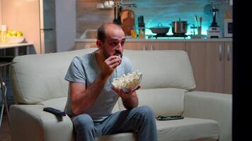 Upset caucasian man watching sport match alone in the living room while hus favourite team looses the game, eating popcorn late at night video