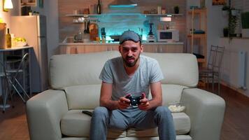 Upset pro gamer sitting in front of television losing soccer games holding wireless controller. Frustrated man with sleep mask lying on sofa late at night in kitchen video