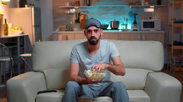 Portrait of man with beard holding popcorn bowl while watching entertainment movie on television. Concentrated person with eye sleep mask sitting on sofa late at night in kitchen video