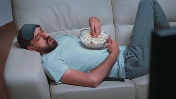 Tired man relaxing on sofa in front of television eating popcorn while watching movie show. Caucasian male with beard looking entertainment series late at night in kitchen video