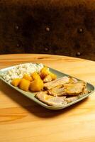 A succulent roasted pork loin sits on a plate alongside golden-brown roasted potatoes and a mound of fluffy white rice. The dish is presented in a cozy restaurant photo