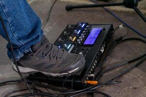 A foot steps on a guitar effects pedal, captured from the side. The shoe is a simple sneaker, and the pedal rests on a textured floor. The focus is on the action, with a sense of anticipation. photo